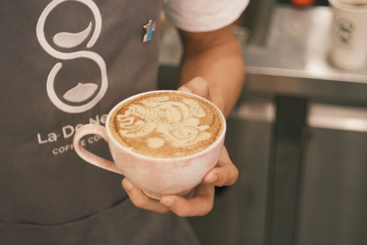 Close-up of a barista holding a cappuccino with intricate latte art, indoors in a coffee shop.