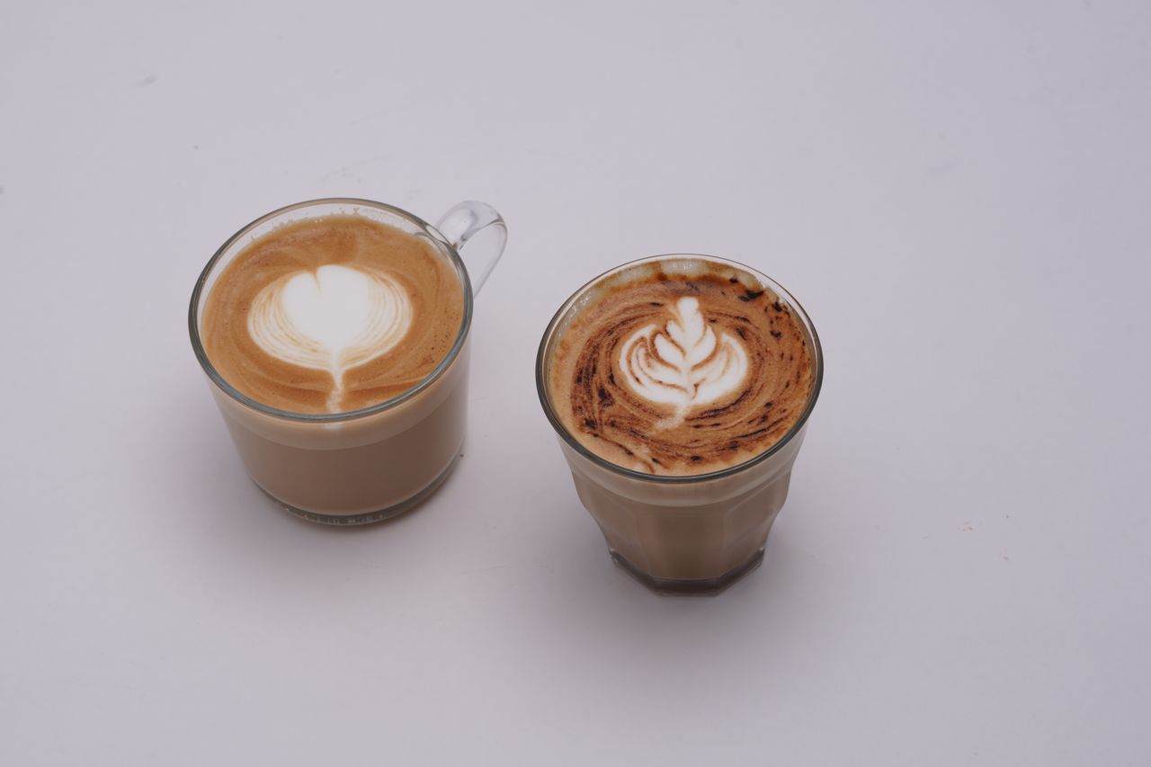 Two glass cups of latte with intricate latte art on white background.
